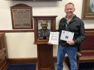 Read more about the article Charles Duncan passing out Part 4 of the Alaska State Bank series in the state capital in front of a bust of Bill Egan who was one of the founders of the State of Alaska.
